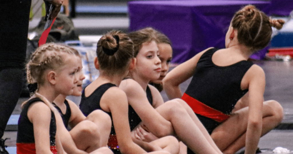 Youth gymnasts sit together between rotations at a meet, representing competitive gymnastics and youth sports culture and team routines in gymnastics levels explained.